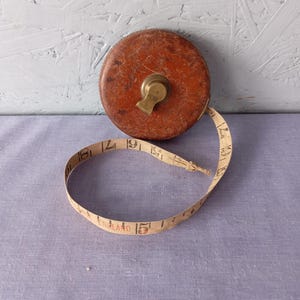 Puede incluir: Un metro de cinta vintage marrón y latón. La caja circular contiene una cinta color crema marcada con números. La cinta se extiende desde la caja, mostrando el número 15. La palabra "ENGLAND" está impresa en la cinta.