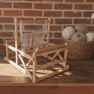 Puede incluir: Un telar de mesa de madera clara que presenta un marco vertical con hilos de urdimbre blancos ensartados a través del lizo. El telar tiene una base resistente con soportes laterales en cruz y componentes de tensión de plástico rojo.