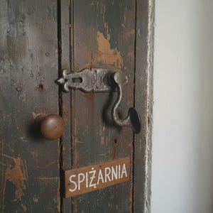 May include: Close-up of a weathered wooden door with a dark, distressed finish. The door features a rustic metal latch and a round wooden knob. A small rectangular sign reads "SPIŻARNIA".