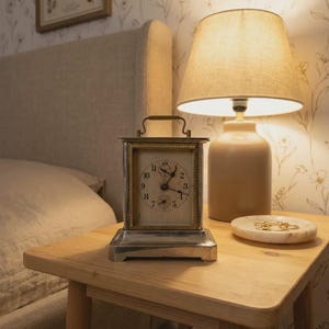 May include: A rectangular silver-toned carriage clock with a white face, black Arabic numerals, and a top handle. The clock features a gold-toned inner frame and sits on a wooden nightstand next to a beige table lamp and a small white marble dish.
