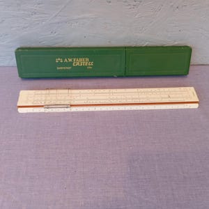 May include: A vintage slide rule with a cream-coloured body and brown accents, next to a green case. The case is labelled "A.W. Faber Castell Darmstadt 1/54". The slide rule is a mathematical tool used for calculations.