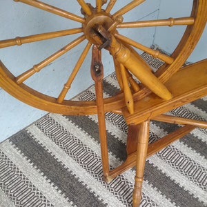 May include: A wooden spinning wheel featuring a large circular frame with multiple spokes radiating from a central hub. The structure includes a vertical support post, a horizontal base, and a connecting rod, all finished in a warm, light brown tone.