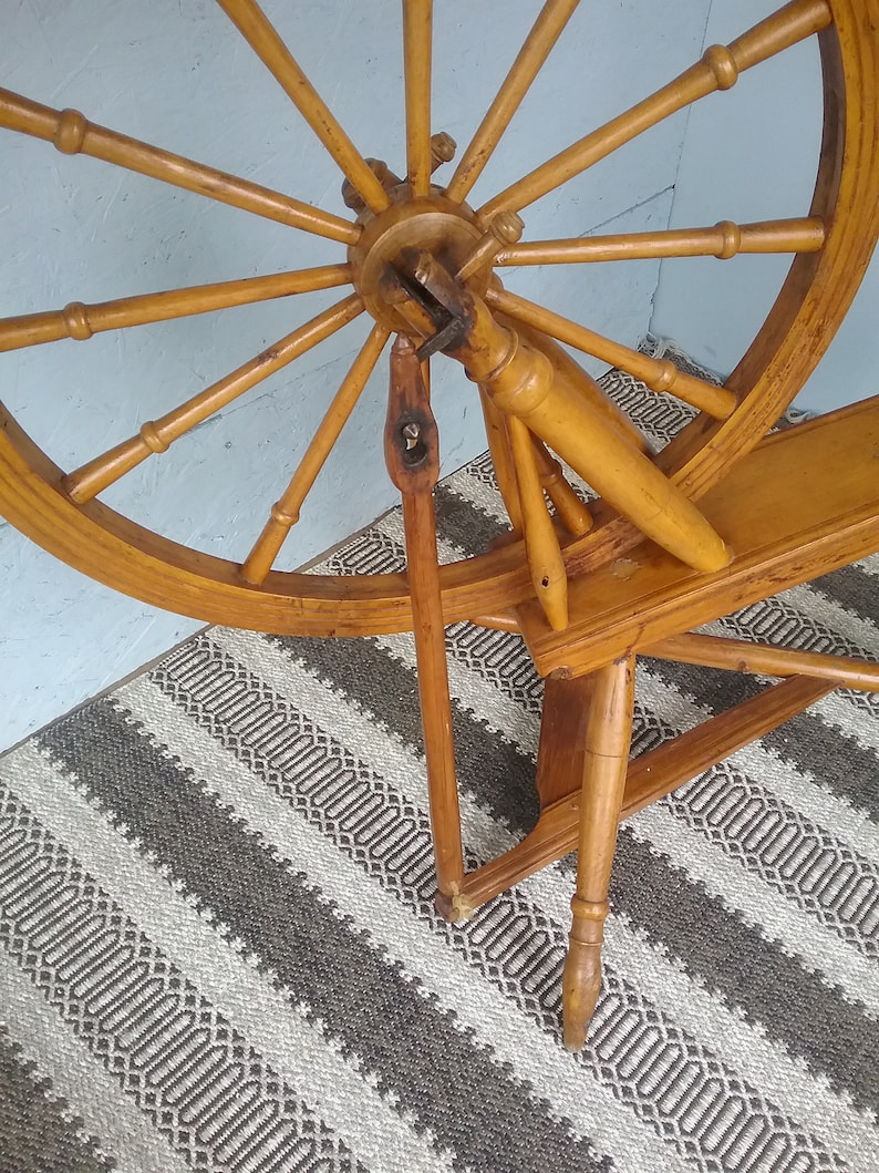 May include: A wooden spinning wheel featuring a large circular frame with multiple spokes radiating from a central hub. The structure includes a vertical support post, a horizontal base, and a connecting rod, all finished in a warm, light brown tone.