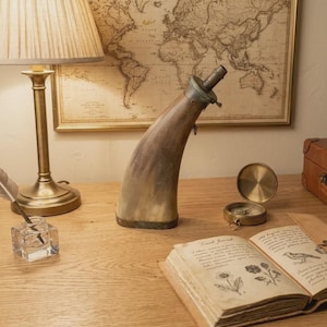Op de afbeelding: Een gebogen, hoornvormige kruitfles met een metalen tuit en voet, geplaatst op een houten oppervlak naast een open reisdagboek met botanische illustraties, een koperen kompas en een glazen inktpot met een ganzenveer.