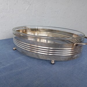 May include: A silver serving dish with a glass insert and a decorative rim. The dish has four small feet and a handle on one side.