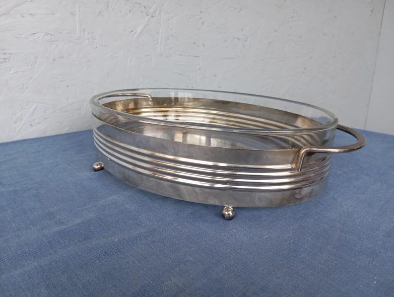 May include: A silver serving dish with a glass insert and a decorative rim. The dish has four small feet and a handle on one side.