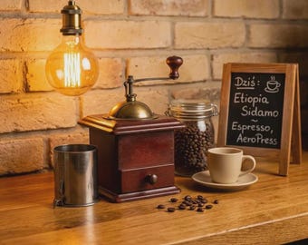 May include: A vintage-style wooden coffee grinder with a brass crank, a clear glass jar filled with coffee beans, a stainless steel cup, and a beige ceramic cup on a saucer. A small chalkboard displays the text Dziś: Etiopia Sidamo Espresso, AeroPress.