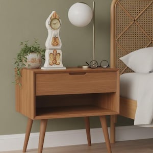 May include: A wooden nightstand with a single drawer and an open shelf, supported by four tapered legs. On top sits a white decorative clock with painted floral motifs, a potted green plant, a spherical lamp, and a pair of black-rimmed glasses.