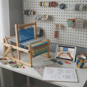 May include: A wooden table loom with a partially woven blue, orange, and yellow textile. The setup includes two small white bowls of colorful beads, a tablet displaying a weaving tutorial, a patterned sketchbook, and a geometric print mug.