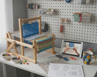 Op de afbeelding: Een houten tafelweefgetouw met een gedeeltelijk geweven blauw, oranje en geel textiel. De set bevat twee kleine witte schaaltjes met gekleurde kralen, een tablet met een weeftutorial, een schetsboek met patronen en een geometrische mok.