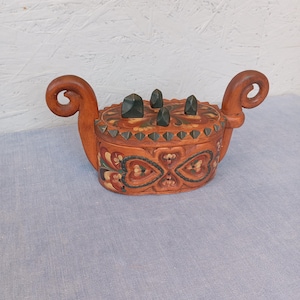 May include: A wooden box with a lid, featuring a carved design and dark green stone accents. The box has a reddish-brown finish with floral and heart-shaped patterns in red, green, and yellow. It has curved handles on each side.