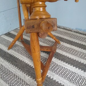 May include: A wooden spinning wheel featuring a light brown finish with a visible grain. The structure includes a horizontal beam with a turned wooden knob, a triangular base with three tapered legs, and a large vertical wheel component.