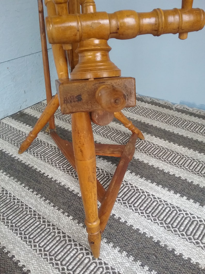 May include: A wooden spinning wheel featuring a light brown finish with a visible grain. The structure includes a horizontal beam with a turned wooden knob, a triangular base with three tapered legs, and a large vertical wheel component.