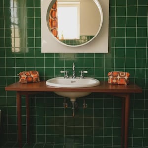 May include: A bathroom vanity featuring a white oval sink set into a dark wood table with four tapered legs. Above the sink is a circular mirror with a white square frame. Two orange and brown patterned pouches sit on the table surface.