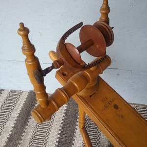 May include: A light brown wooden spinning wheel featuring a flyer assembly with a bobbin and a whorl. The horizontal wooden frame is stamped with the text APS HJULBACK. The structure includes turned wooden uprights and a central spindle mechanism.