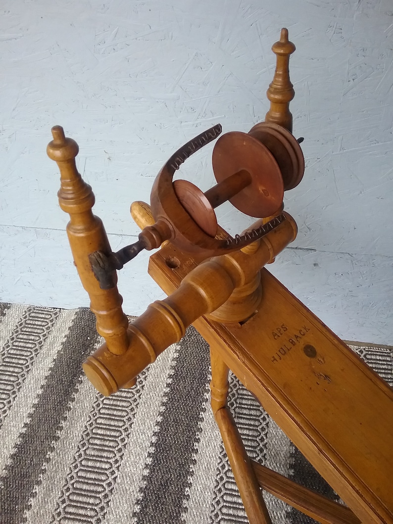 May include: A light brown wooden spinning wheel featuring a flyer assembly with a bobbin and a whorl. The horizontal wooden frame is stamped with the text APS HJULBACK. The structure includes turned wooden uprights and a central spindle mechanism.
