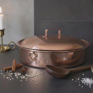 Op de afbeelding: Een ronde koperen pan met bijpassend deksel, voorzien van twee ringvormige handgrepen aan de zijkanten en een handgreep op het deksel. De pan wordt vergezeld door een houten lepel, een kommetje zout, kaneelstokjes, steranijs en rijstkorrels.