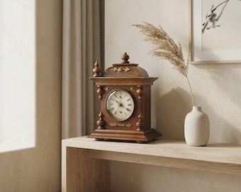 May include: A brown wooden mantel clock with a circular face, Roman numerals, and gold-toned metal accents. The clock features a decorative top finial and a structured base. Beside it sits a white ceramic vase containing a single dried pampas grass stem.