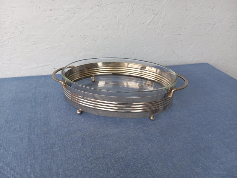May include: Silver serving dish with a glass insert and three feet. The dish has two handles.