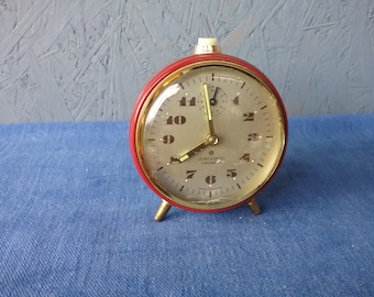May include: A red alarm clock with gold accents and a white face. The clock has black numbers and hands. The clock face says "Junghans  Cavalier".