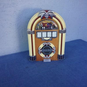 May include: A vintage-style, red, white, and brown tabletop jukebox with a palm tree design. The jukebox has a clear plastic window and a silver speaker grill.