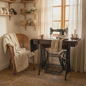 Può includere: Una macchina da cucire a pedale Husqvarna nera vintage si trova su un tavolo in legno con un tessuto a maglia beige drappeggiato sopra. Accanto al tavolo c'è una sedia in vimini coperta da una coperta color crema e scaffali in legno con filati e cesti.