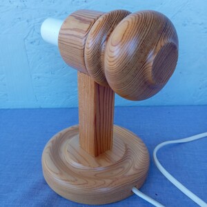 Op de afbeelding: Een houten tafellamp met een ronde basis en een cilindrische steel. De lampenkap is eenvoudig, afgerond en gemaakt van hout. De lamp heeft een wit snoer.
