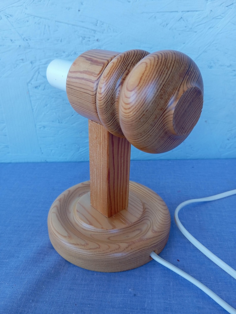 Op de afbeelding: Een houten tafellamp met een ronde basis en een cilindrische steel. De lampenkap is eenvoudig, afgerond en gemaakt van hout. De lamp heeft een wit snoer.