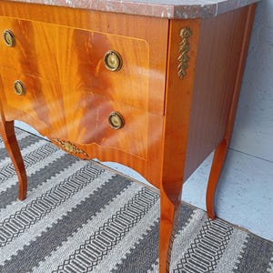 May include: A wooden chest of drawers with a marble top. The chest has three drawers with brass knobs and decorative gold accents. The wood has a warm, reddish-brown finish and curved legs. The marble top has a pinkish hue.