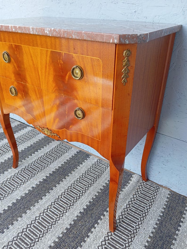 May include: A wooden chest of drawers with a marble top. The chest has three drawers with brass knobs and decorative gold accents. The wood has a warm, reddish-brown finish and curved legs. The marble top has a pinkish hue.