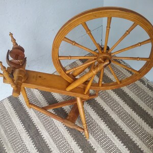 May include: A light brown wooden spinning wheel featuring a large circular wheel with twelve spokes, a horizontal base, and a flyer assembly with a bobbin. The wood has a smooth finish and the structure is supported by three legs on a flat surface.