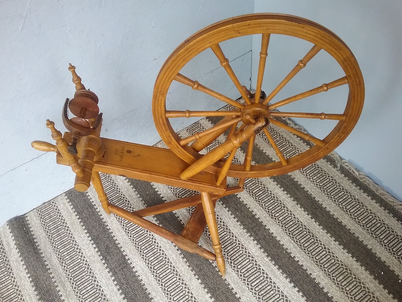 May include: A light brown wooden spinning wheel featuring a large circular wheel with twelve spokes, a horizontal base, and a flyer assembly with a bobbin. The wood has a smooth finish and the structure is supported by three legs on a flat surface.