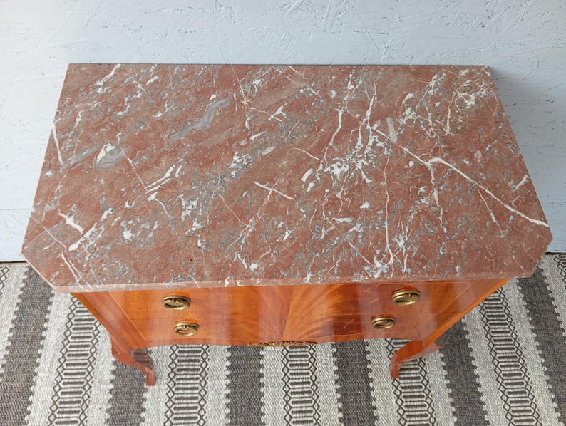 May include: A wooden chest of drawers with a red marble top. The chest has two drawers with brass handles. The wood has a warm, reddish-brown tone, and the marble top features white veining.