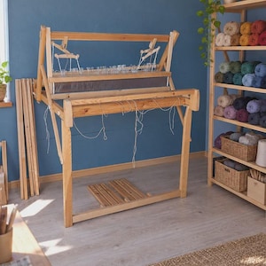 Op de afbeelding: Een groot houten weefgetouw met een natuurlijke lichthouten afwerking, voorzien van een horizontaal frame, een riet, schachten en een voetpedaalmechanisme aan de basis. Het weefgetouw heeft verticale steunen en losse witte draden.