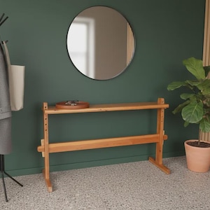Puede incluir: Escena interior moderna con una consola de madera, un espejo redondo y un perchero. Un abrigo gris y un bolso tote cuelgan del perchero. Una planta en maceta está cerca. La habitación tiene una pared verde.