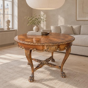 Op de afbeelding: Een ronde houten tafel met een gepolijst bruin fineerblad met een stervormig patroon. De tafel heeft gebogen poten met sierlijk houtsnijwerk en klauwpoten, verbonden door een centrale kruisvormige dwarsbalk met decoratieve details.