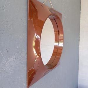 May include: A square wall mirror with a glossy, deep brown frame featuring a circular center cutout. The frame has a smooth, reflective surface that curves inward toward the round glass mirror. A thin, light-colored cord is attached for hanging.