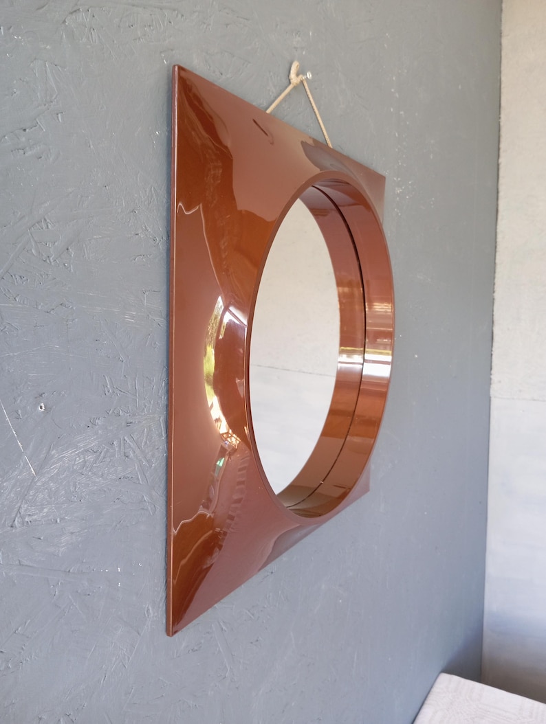 May include: A square wall mirror with a glossy, deep brown frame featuring a circular center cutout. The frame has a smooth, reflective surface that curves inward toward the round glass mirror. A thin, light-colored cord is attached for hanging.