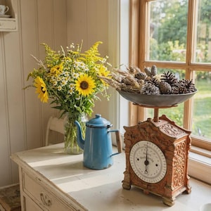 Op de afbeelding: Een boeket gele zonnebloemen en witte madeliefjes in een glazen pot, een lichtblauwe emaille koffiepot en een sierlijke, verweerde oranje metalen keukenweegschaal met een schaal vol dennenappels en gedroogde tarwe op een wit oppervlak.