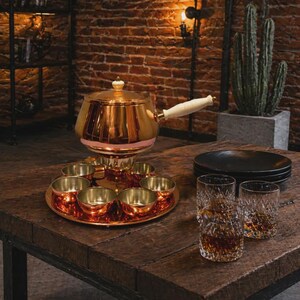 Puede incluir: Un juego de fondue de cobre con una bandeja y seis cuencos pequeños. El juego incluye una olla con tapa y asa blanca. Dos vasos de cristal y platos negros sobre una mesa de madera.