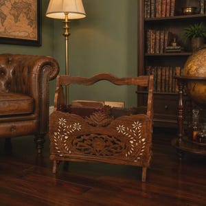 May include: A wooden magazine rack with intricate floral and starburst cutouts. The rack is a warm brown color and has a curved handle. Books are visible inside the rack. The rack is in a room with a leather chair and a floor lamp.