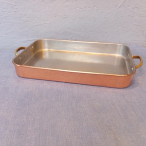 May include: A rectangular copper-coloured baking pan with a silver interior. The pan has two brass-coloured handles on the sides. The pan is on a blue surface.