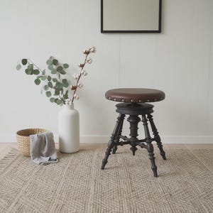 May include: A brown leather-topped stool with a dark wooden base. The stool has a round seat with decorative studs around the edge. The base features ornate, turned legs. A white vase with greenery and a woven basket are in the background.