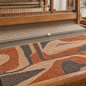Può includere: Un tappeto tessuto con un motivo astratto di forme organiche in arancione bruciato, grigio antracite e beige. Il tappeto è in fase di produzione su un telaio in legno, con i fili di ordito visibili e il pettine in metallo posizionato.