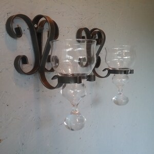 May include: A black metal wall-mounted candle holder with three glass candle holders. The candle holders are clear glass with a teardrop shape.