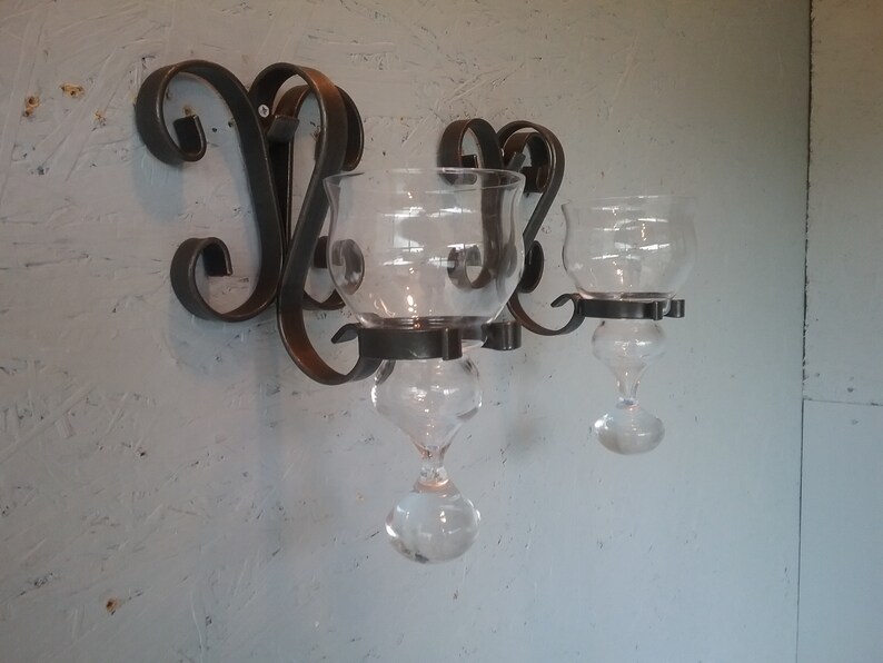 May include: A black metal wall-mounted candle holder with three glass candle holders. The candle holders are clear glass with a teardrop shape.