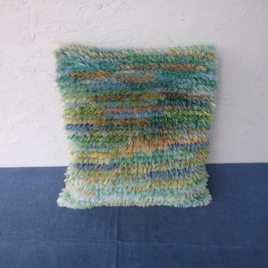 May include: A square, textured pillow with a shaggy, multi-colored design. The pillow features a blend of blue, green, and orange hues, creating a vibrant and tactile appearance. The pillow is placed on a blue surface.
