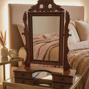 May include: A rectangular wooden vanity mirror with a swivel frame and ornate carved detailing at the top. The base features two small drawers on either side of a central arched cutout, providing a compact storage solution for small items.