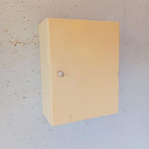 May include: A rectangular, cream-colored metal cabinet with a single door and a small, round silver handle. The cabinet is mounted on a light blue wall. The cabinet appears to be empty.