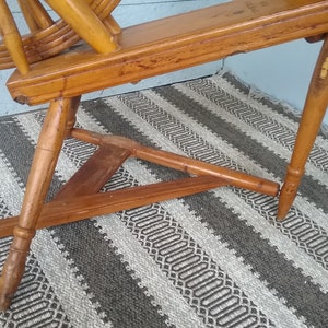May include: A vintage wooden spinning wheel featuring a natural light brown finish with visible wood grain. The structure includes a large circular wheel, a horizontal frame, and three tapered legs connected by a triangular wooden base support.
